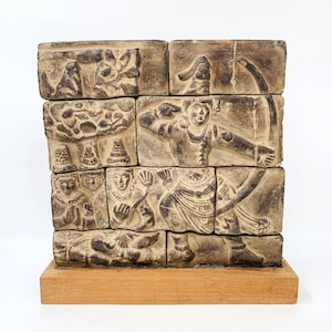 Op de afbeelding: Een gestapelde sculptuur bestaande uit rechthoekige blokken, elk met gedetailleerde reliëfsnijwerken. De snijwerken tonen figuren in verschillende poses, waaronder een boogschutter met een getrokken boog. De sculptuur is gemonteerd op een houten voet, met oude kunst.
