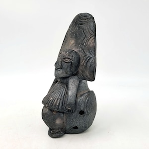 May include: Black ceramic figurine of a seated figure with a large, patterned headdress. The figure has a stylized face and is wearing a skirt-like garment.