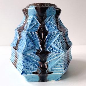 Op de afbeelding: Een keramische vaas met een geometrisch ontwerp. De vaas heeft een lichtblauwe gespikkelde glazuur met donkerbruine accenten. De vaas heeft een zeshoekige vorm met een getextureerd oppervlak en een uitlopende bovenkant.