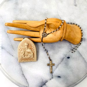 May include: A white and gold rosary with clear glass beads and a small metal heart charm. The rosary is resting on a wooden hand. A white ceramic pendant with a relief image of Jesus Christ is also pictured.