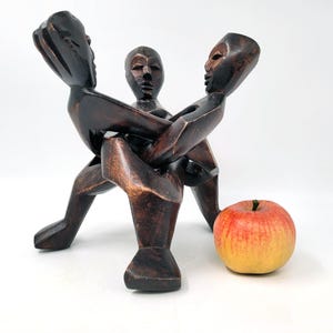 May include: A dark brown wooden sculpture depicting three figures intertwined. The sculpture has a smooth, polished finish. A red and yellow apple sits next to the sculpture. The sculpture is approximately 10 inches tall.