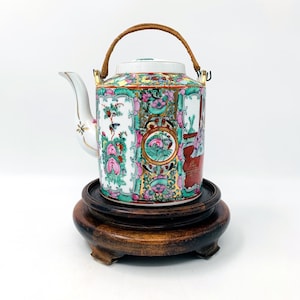 May include: A decorative porcelain teapot with a woven handle, featuring a colourful floral design with pink, green, and gold accents. The teapot is displayed on a dark wooden stand, showcasing its intricate details and craftsmanship.