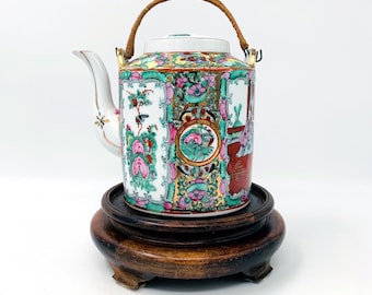 May include: A decorative porcelain teapot with a woven handle, featuring a colorful floral design with pink, green, and gold accents. The teapot is displayed on a dark wooden stand, showcasing its intricate details and craftsmanship.