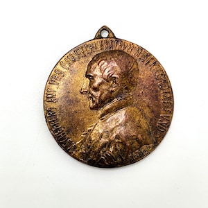 Puede incluir: Medalla redonda de color bronce con el retrato de perfil de un hombre. La medalla tiene un lazo en la parte superior para colgarla y una inscripción alrededor del borde. El rostro del hombre es detallado y la medalla tiene un aspecto envejecido.
