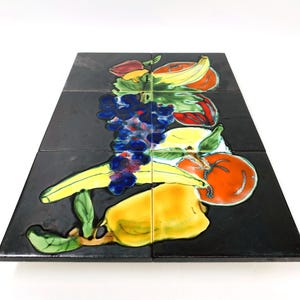 May include: A set of six black ceramic tiles arranged to form a square. Each tile features a colorful illustration of various fruits, including bananas, grapes, and a tomato. The fruits are painted in vibrant shades of yellow, red, and green.