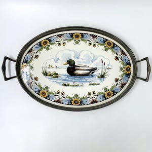 Puede incluir: Una bandeja ovalada para servir con un borde plateado y un centro de cerámica blanca. El centro de cerámica presenta un diseño pintado a mano de un pato nadando en un estanque con agua azul y verde. El borde de la bandeja está decorado con un patrón floral en tonos azules, verdes, amarillos y rojos.