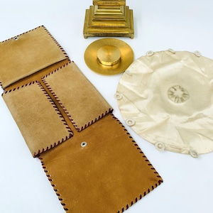 May include: A tan leather wallet with dark brown stitching is open, revealing multiple compartments. A gold-colored decorative object and a cream-colored fabric item are also present. The wallet is made of suede-like material.