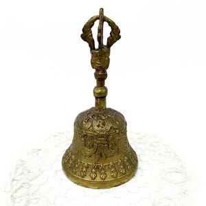 May include: A vintage brass bell with detailed embossed patterns. The bell has an ornate handle with a decorative top. The bell is a warm gold colour and rests on a white lace surface. The bell is likely used for religious or decorative purposes.