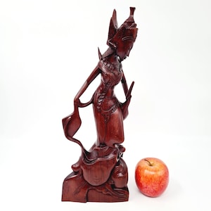 以下が含まれることがあります： 伝統的な衣装を着た女性の彫刻。動物が彫られた台座の上に立っています。彫刻は濃い赤色の木で作られており、高さは約30センチメートルです。