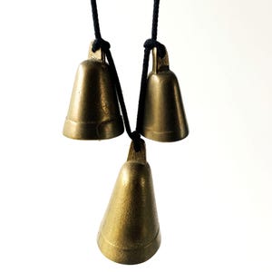 May include: Three brass-coloured metal bells of varying sizes suspended from a single black cord. Each bell features a smooth, conical shape with a small loop at the top for attachment. The metal has a muted, metallic gold finish with subtle variations.