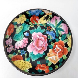 Op de afbeelding: Een ronde, decoratieve schotel met een zwarte achtergrond en een bloemenontwerp. De bloemen zijn in roze, rood, geel en blauw, met groene bladeren. De schotel heeft een donkerbruine rand.