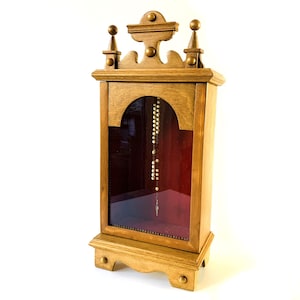 Puede incluir: Un expositor de madera con frente de cristal e interior de terciopelo rojo. Un rosario cuelga dentro del expositor. El expositor tiene una parte superior e inferior decorativas y está hecho de madera clara.