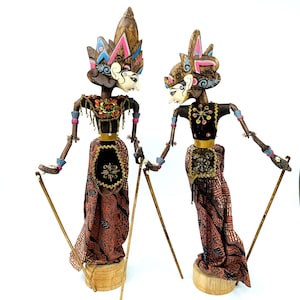 Puede incluir: Dos marionetas tradicionales indonesias Wayang Golek. Las marionetas tienen cabezas de madera tallada, tocados ornamentados y trajes detallados en tonos marrón, negro, rosa y dorado. Cada marioneta sostiene una fina varilla de madera.