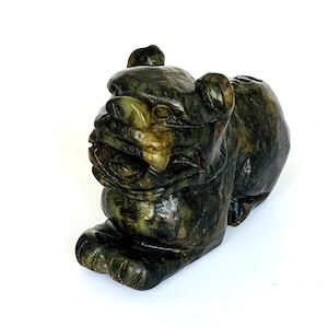 Op de afbeelding: Een gebeeldhouwd stenen beeldje van een mythisch wezen, mogelijk een Foo Dog. De sculptuur is voornamelijk donkergroen met lichtere groene en gele accenten. Het wezen zit in een zittende positie met een felle uitdrukking.