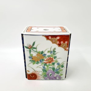 May include: A white ceramic square box with a floral design. The box has a red, orange, yellow, blue, and green floral pattern. The top of the box has a small hole in the center.