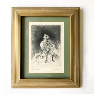 Op de afbeelding: Een ingelijste zwart-wit ets van een haan met een gedetailleerde gouden lijst. Het kunstwerk toont een haan met een gedetailleerde staart en een klein dier op de achtergrond. Het kunstwerk is geplaatst tegen een groene passe-partout.
