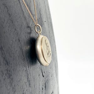 Op de afbeelding: Een zilverkleurige ovale medaillonhanger aan een fijne ketting. Het medaillon heeft een gedetailleerde reliëf afbeelding op de voorkant. De ketting en het medaillon worden getoond tegen een donker houten oppervlak.