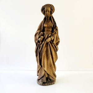 Op de afbeelding: Een houten beeld van een geklede figuur, mogelijk religieus, met een serene uitdrukking. De figuur houdt een voorwerp vast en is gehuld in vloeiende kleding. Het beeld heeft een verweerde, verouderde uitstraling, wat suggereert dat het antiek is.