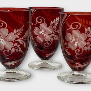 Puede incluir: Tres copas de vino de color rojo rubí con diseños florales grabados. Cada copa presenta una flor grande y delicados patrones de hojas. Las copas tienen una base clara y redondeada y una parte superior acampanada.