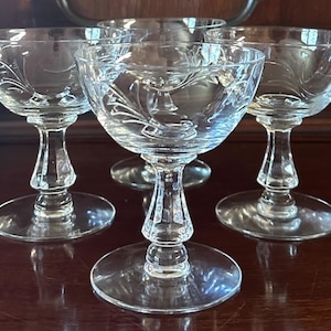 Może przedstawiać: Zestaw czterech przezroczystych kieliszków z wytrawionymi wzorami kwiatowymi. Kieliszki mają zaokrągloną czarkę, dekoracyjną nóżkę i okrągłą podstawę. Ustawione na ciemnej, odbijającej światło powierzchni.