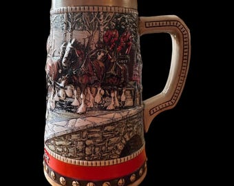 May include: A ceramic beer stein featuring a detailed relief scene of horses pulling a carriage over a stone bridge. The stein has a beige top and handle, a red band, and a decorative base. A collectible item.