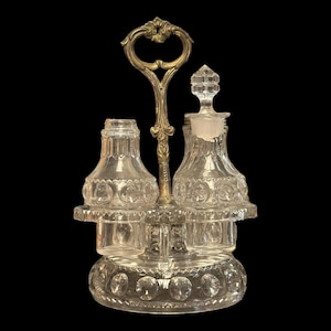 May include: An antique clear glass condiment set with a decorative silver-toned handle. The set includes two bottles and two small containers, all resting on a round, patterned base. The bottles have decorative stoppers.