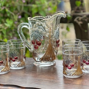 May include: A set of six clear glass cups and a jug with a cherry and leaf design. The jug has a gold trim and the cups have a gold rim.