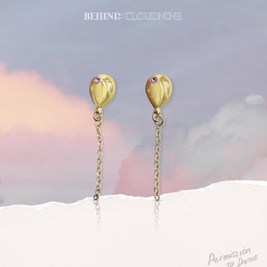 May include: A pair of gold balloon earrings with a pink gemstone accent. The earrings dangle from a delicate gold chain.