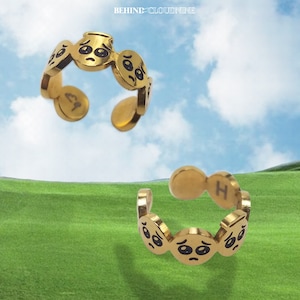 May include: Two gold adjustable rings with a design of multiple sad faces. The rings are on a green grassy surface with a blue sky in the background.
