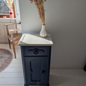Peut inclure: Une petite table de chevet bleu foncé avec un dessus en marbre blanc. La table de chevet a un seul tiroir et une porte de placard avec un design décoratif. La table de chevet est posée sur un sol en bois blanc.