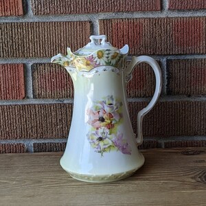 May include: A white porcelain jug with a floral design. The jug has a lid with a floral design and a handle. The jug is decorated with pink, white, and yellow flowers.
