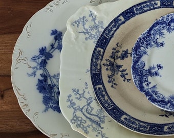 Op de afbeelding: Een verzameling antieke wit en blauw porseleinen borden met bloemen- en vogelontwerpen. De borden variëren in grootte en vorm, met gouden rand op één. De borden zijn gerangschikt op een houten oppervlak.