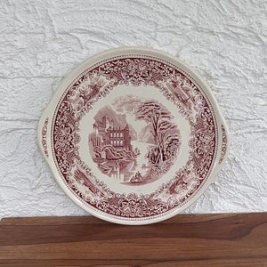 Puede incluir: Un plato de servir de cerámica blanca con un diseño floral rosa y una escena de un castillo junto a un lago. El plato tiene dos asas a los lados.