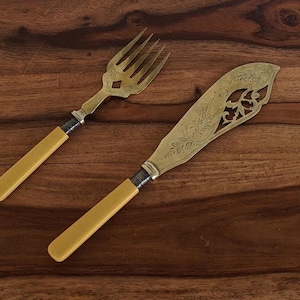 Puede incluir: Un juego de cubiertos vintage con mangos de plástico amarillo. El tenedor y el cuchillo tienen diseños ornamentados y están hechos de plata.