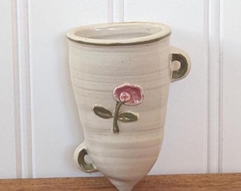 May include: White ceramic wall vase with a green and pink flower design. The vase has a rounded bottom and two handles.
