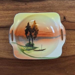 Könnte beinhalten: Eine quadratische Vintage-Keramikschale mit handgemalter Landschaftsszene. Das Design zeigt einen Sonnenuntergang über einem See mit Bäumen und einem kleinen Boot. Die Schale hat einen gelben Rand und zwei Griffe.