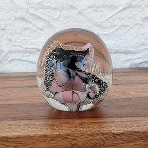 May include: A glass paperweight featuring a pink and black floral design. The paperweight is round and clear, with the floral design visible through the glass.