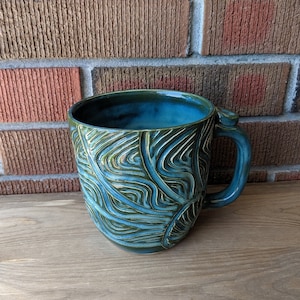 May include: A teal blue ceramic mug with a green and white leafy pattern. The mug has a rounded body and a handle.