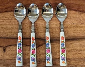 Op de afbeelding: Vier roestvrijstalen lepels met witte handvatten versierd met een bloemenpatroon in roze, blauw en oranje. De lepels zijn gerangschikt op een houten oppervlak.