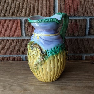 May include: A vintage ceramic pitcher with a yellow, green, and purple glaze. The pitcher has a handle and a detailed design of a face and leaves.