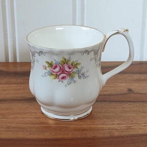 Puede incluir: Taza de té de cerámica blanca con un diseño floral de rosas rosas, verdes y azules. La taza tiene un borde y un asa dorados. Está sobre una superficie de madera. Diseño clásico.