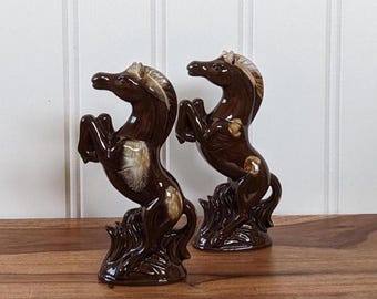 May include: Two brown ceramic horse figurines in a rearing pose. The horses have a glossy finish with lighter brown and beige accents on their manes and bodies. They are mounted on a base that resembles stylized waves.