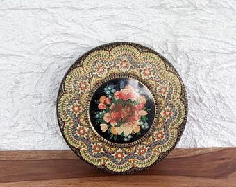 May include: A round, decorative plate with a black background and a colorful floral design. The plate has a gold and green border with a intricate pattern.