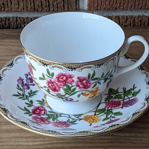 Könnte beinhalten: Ein weißes Tee-Set mit einem Blumenmuster in Rosa-, Gelb- und Grüntönen. Tasse und Untertasse haben einen goldenen Rand.