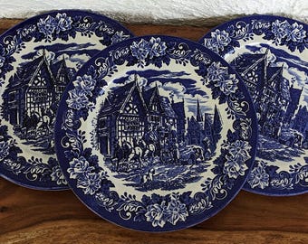 May include: Three blue and white ceramic plates with a floral border and a scene of a village with houses and people. The plates are on a wooden surface.
