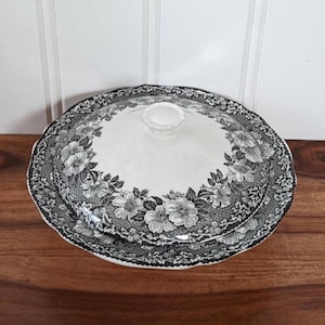 Puede incluir: Una fuente para servir con estampado floral blanco y negro con tapa. El plato tiene un borde festoneado y un pomo transparente en la tapa. El diseño floral se repite alrededor del borde del plato y en la tapa.