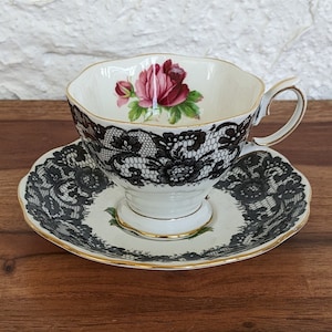 Puede incluir: Una taza de té y un platillo blancos con un diseño de encaje negro y un patrón floral de rosa rosa. La taza de té tiene un borde dorado.
