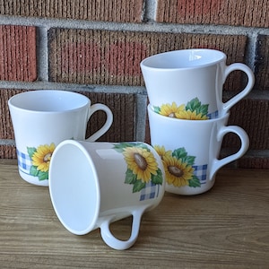 May include: Four white ceramic mugs with a sunflower and blue and white gingham pattern. The mugs are stacked on top of each other.