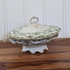 May include: A porcelain serving dish with a lid, featuring a green and gold floral design on a white background. The dish has a handle and sits on a scalloped base. The dish is oval-shaped and rests on a wooden surface.