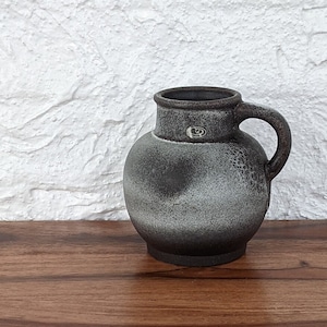 May include: A dark grey ceramic jug with a rounded body and a handle. The jug has a speckled texture and a lighter grey area on the front. A small emblem is visible near the handle. The jug sits on a wooden surface.